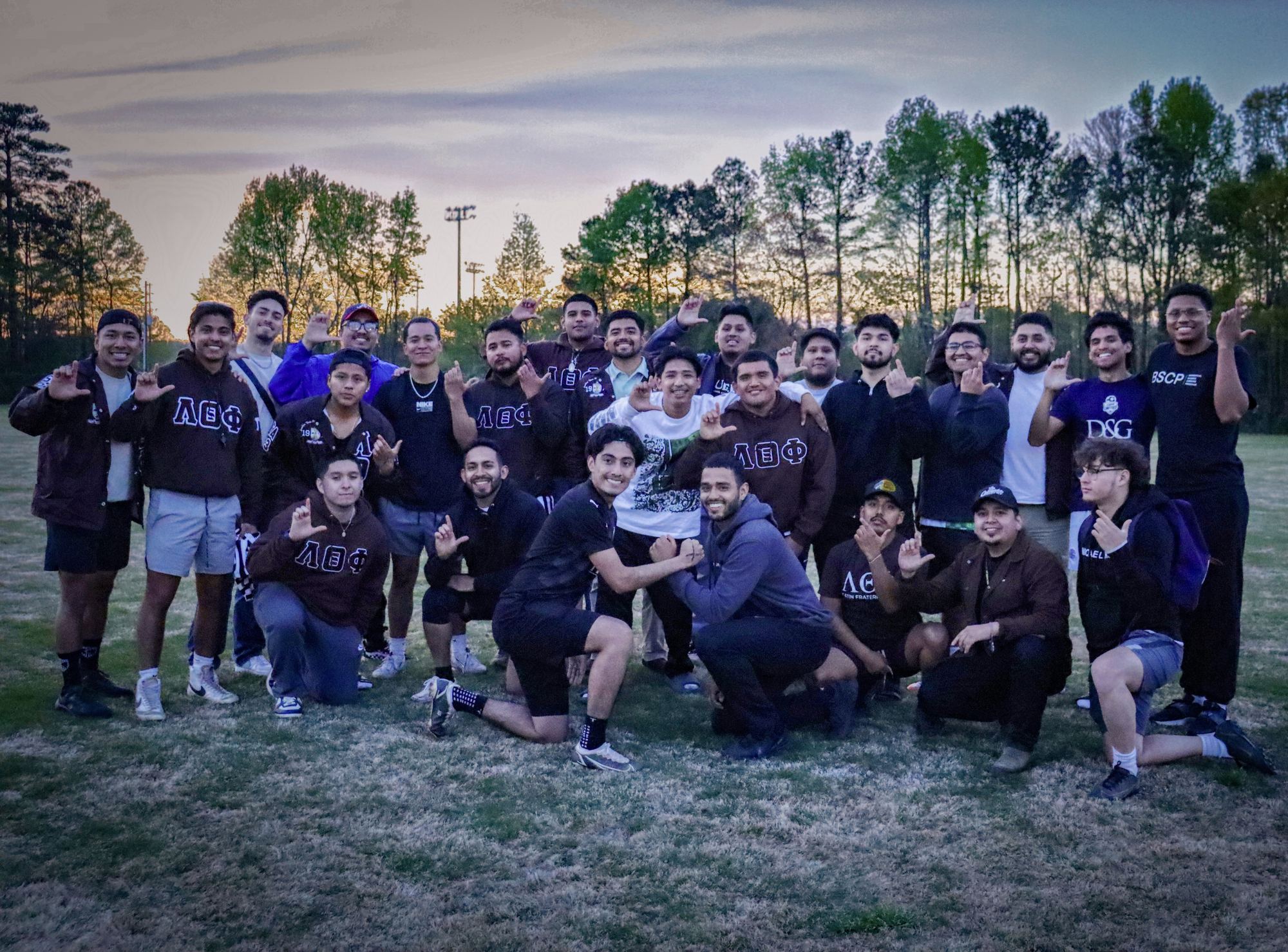 Around twenty brothers gathered on a playing field at sunset, throwing the fraternity's hand sign.