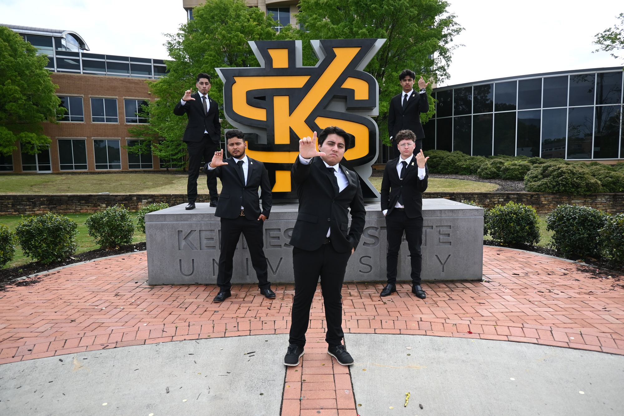 Five brothers in black suits around the Kennesaw State University monument sign.