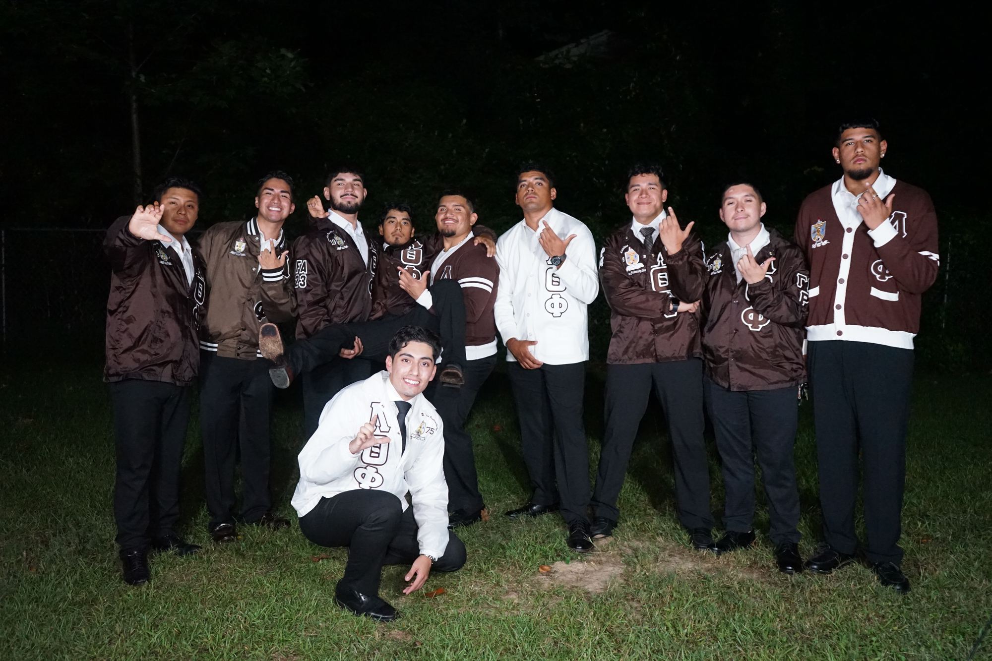 Nine brothers in brown fraternity jackets outdoors at night, throwing the hand sign.