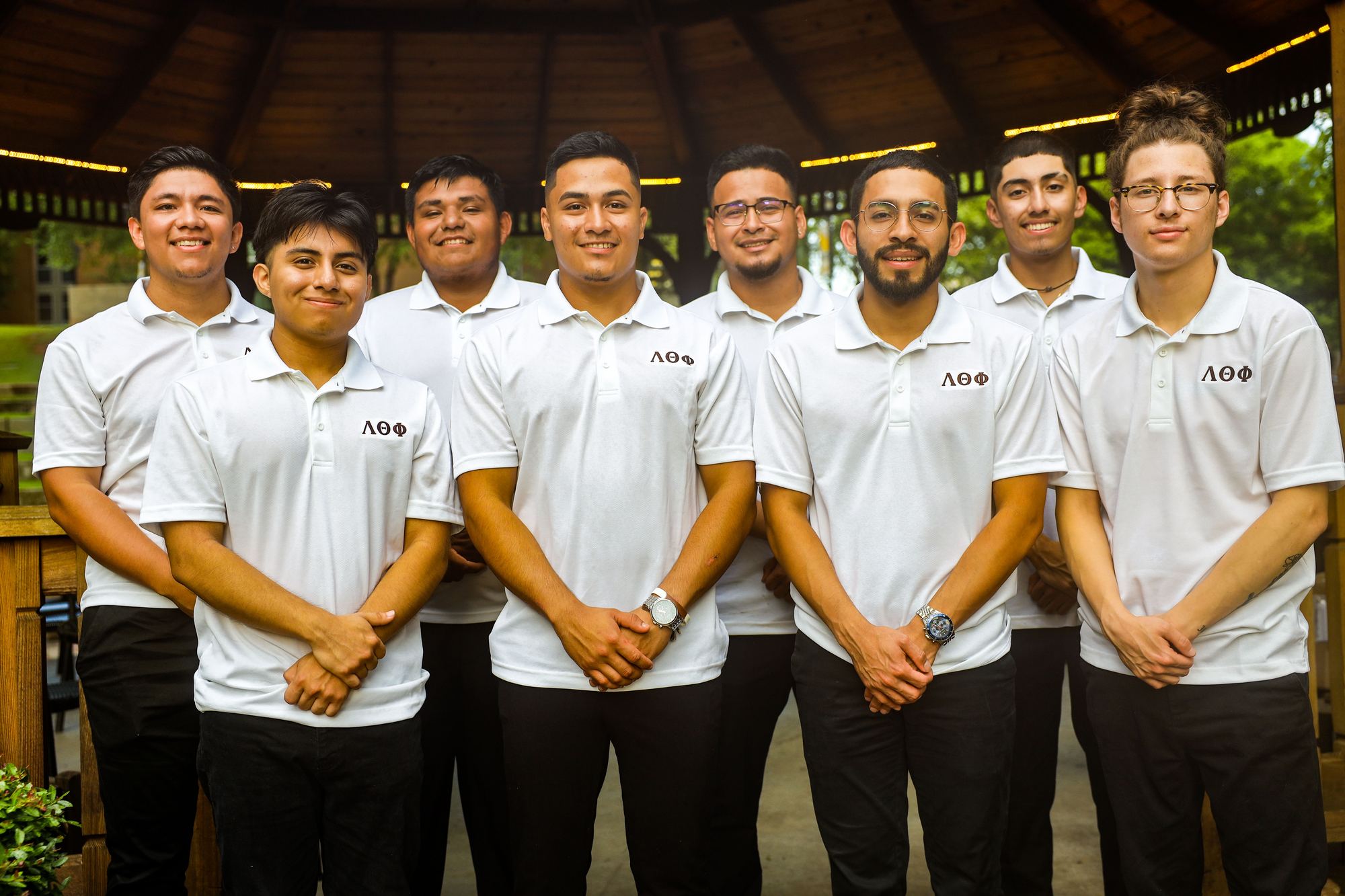 Eight brothers in white Lambda Theta Phi polos standing together under a wooden gazebo.