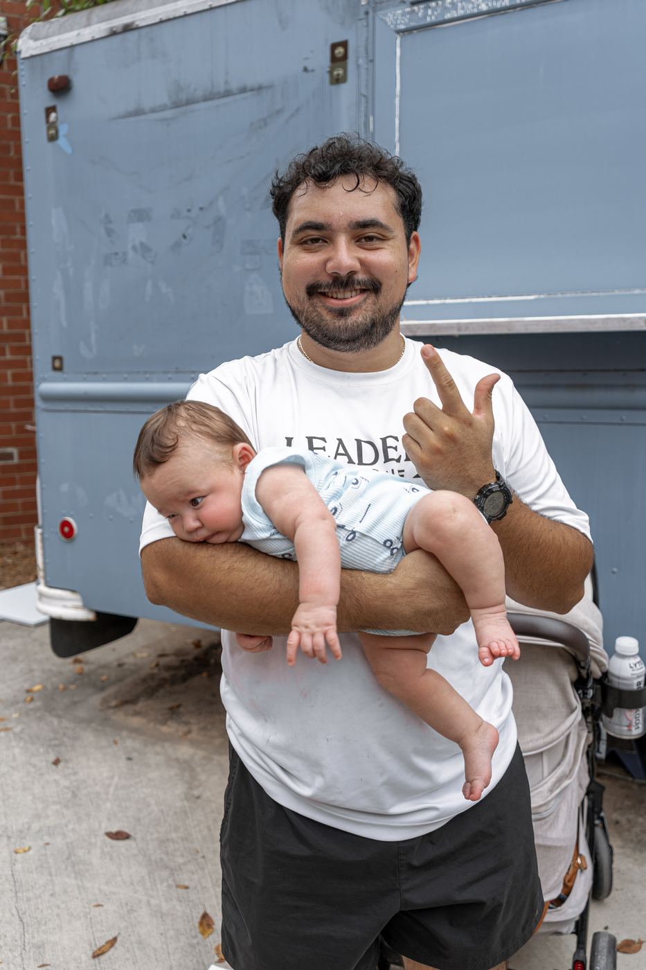 A brother holding a baby in one arm and throwing the fraternity hand sign with the other.