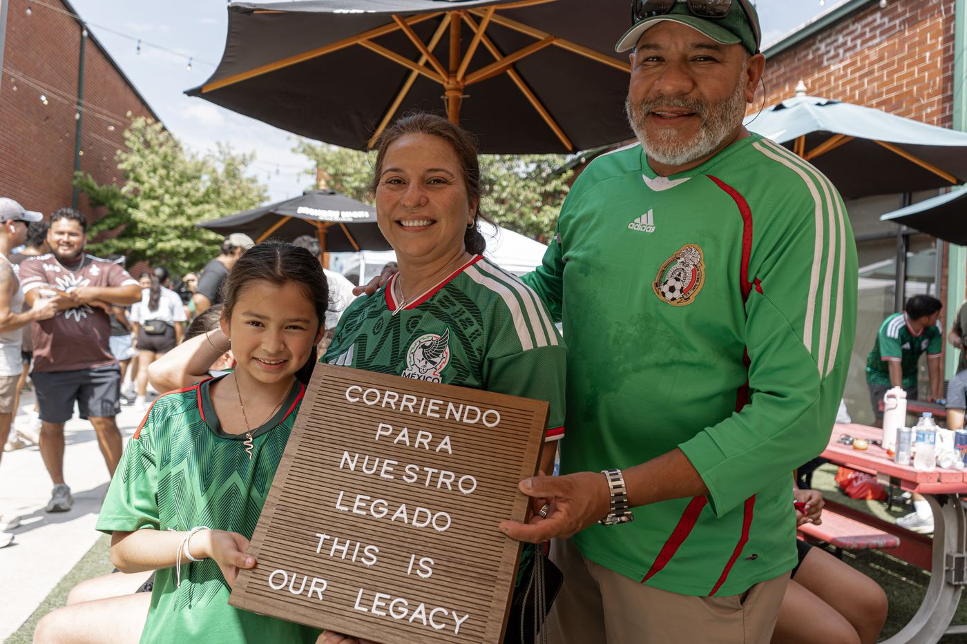 A family of three in Mexico jerseys holding the letterboard together under a patio umbrella.