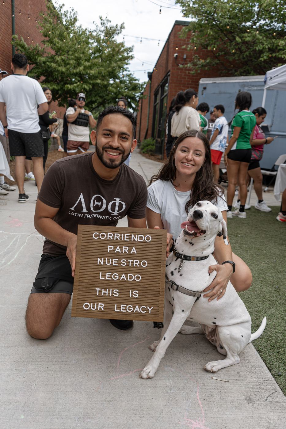 Two people kneeling on the sidewalk with a dalmatian, holding a letterboard reading Corriendo para Nuestro Legado, this is our legacy.