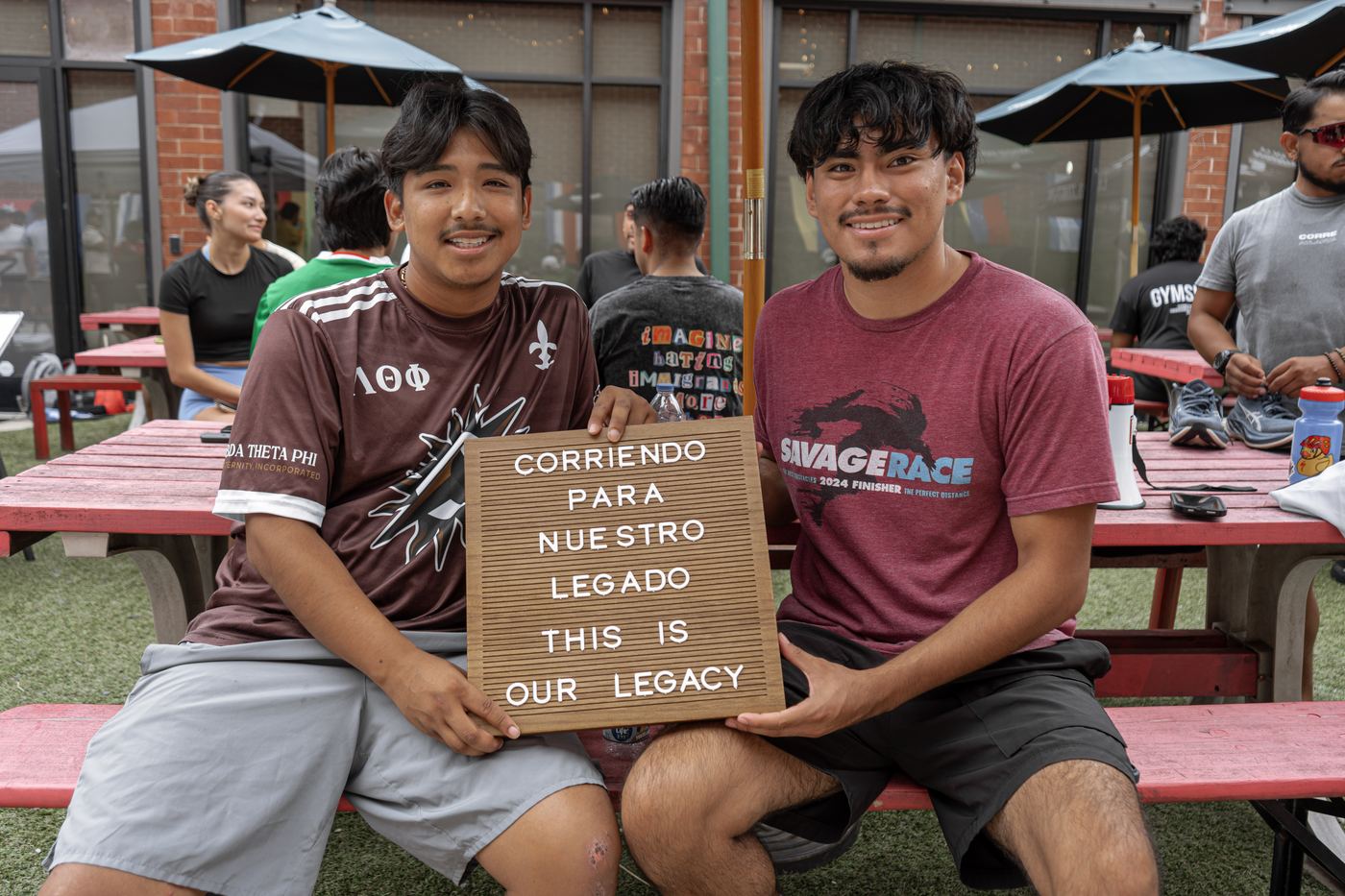 Two runners sitting at a red picnic table holding the Corriendo para Nuestro Legado letterboard.