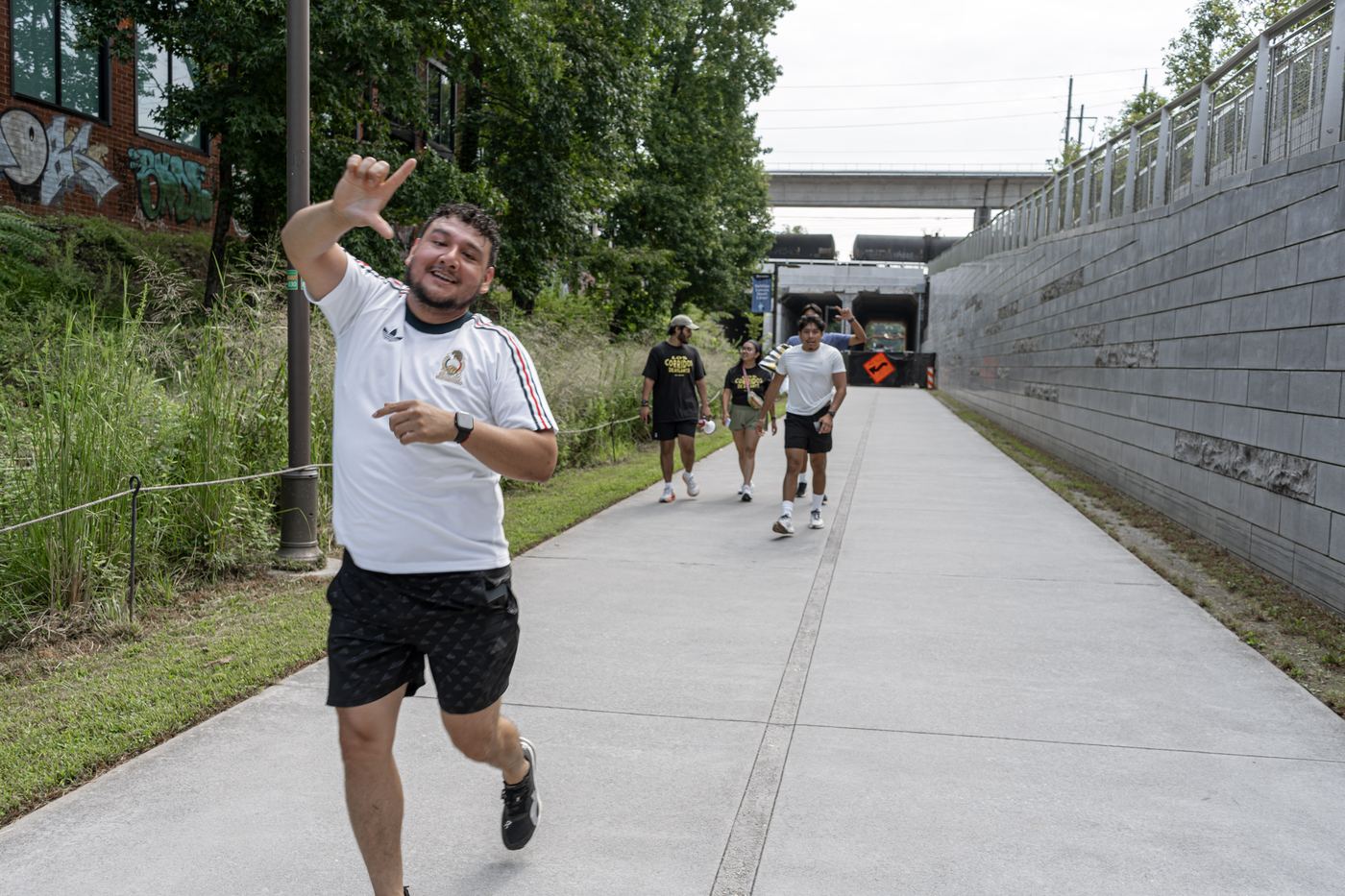 A runner in a white Mexico jersey coming toward the camera throwing the fraternity hand sign, with more runners behind.