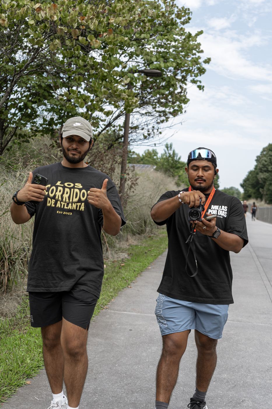 Two runners on the trail, one giving a thumbs up in a Los Corridos de Atlanta shirt, the other checking a camera in a Millas Por Dias shirt.